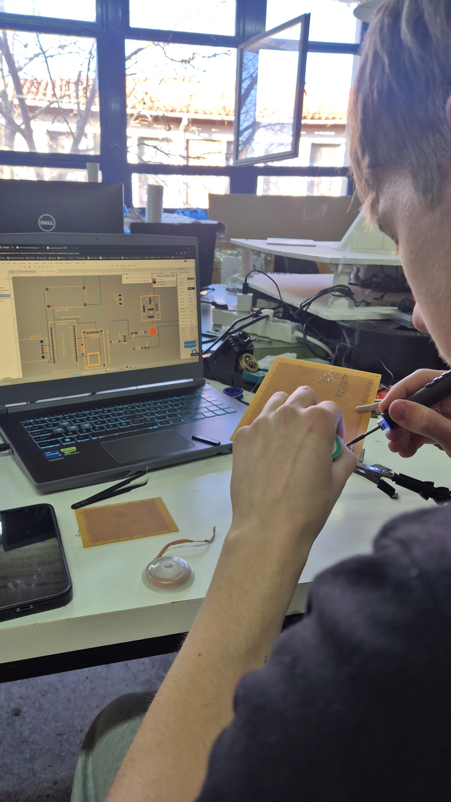 Soldering an electronic circuit board in the workshop beside a laptop displaying a circuit design